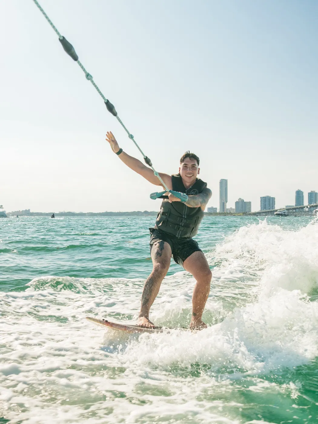 A smiling wakesurfer holding a tow rope with the Miami waterfront in the background.